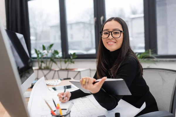 Smiling asian designer holding notebook near blueprint and devices in office — Stock Photo