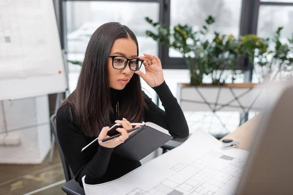 Young asian interior designer in eyeglasses holding notebook near computer and blueprint in office — Stockfoto
