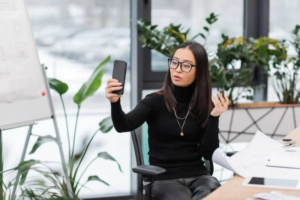 Brunette asian designer in eyeglasses having video call on smartphone near blueprints in office — Stock Photo