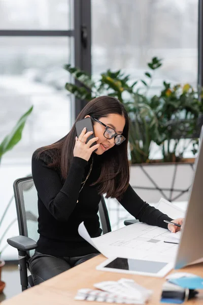 Young asian designer talking on smartphone near blueprint and devices in office — Foto stock
