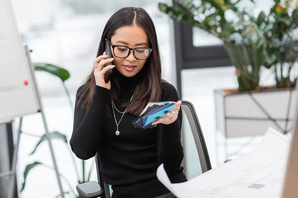 Smiling asian designer talking on smartphone and holding color samples near blueprint in office — Foto stock