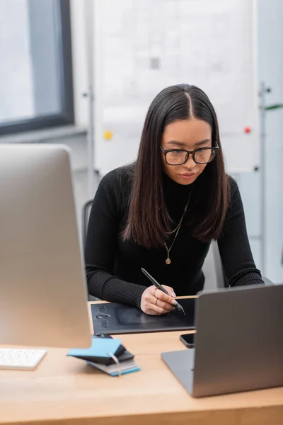 Азиатский дизайнер интерьеров с помощью графического планшета возле ноутбука и компьютера в офисе — стоковое фото