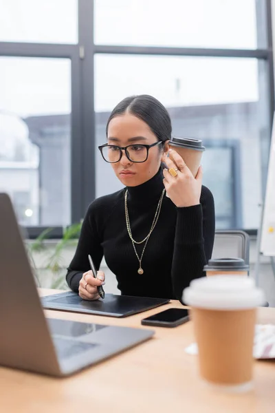 Азиатский дизайнер в очках держит бумажную чашку возле графического планшета и ноутбука в офисе — стоковое фото