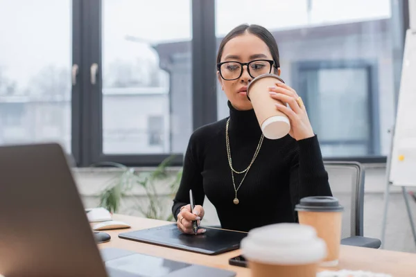 Asian designer holding coffee to go while using graphic tablet in office — Stockfoto