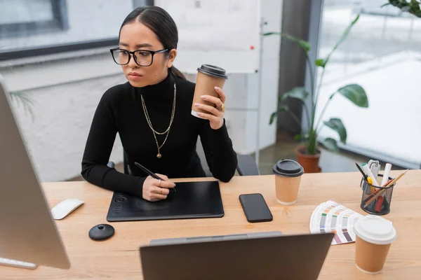 Молодой азиатский дизайнер держит кофе, чтобы идти рядом графические планшеты и гаджеты в офисе — стоковое фото