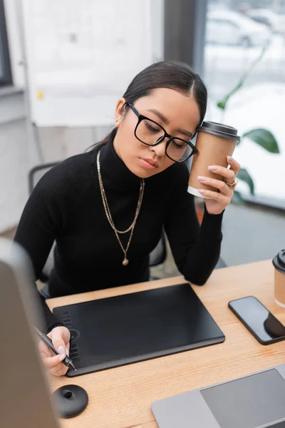 Tired asian interior designer holding coffee to go near devices and graphic tablet in studio — Stock Photo