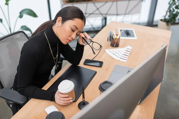 Tired asian interior designer holding eyeglasses and coffee to go in studio — Stock Photo