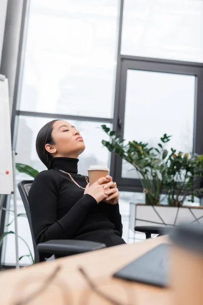 Asian designer holding coffee to go and relaxing near working table in office — Stock Photo