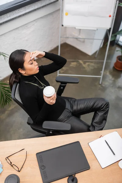 Overhead view of asian designer holding coffee to go while suffering from headache in studio — Stock Photo
