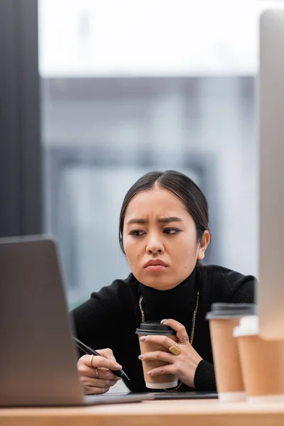 Недовольный азиатский дизайнер держит кофе рядом графические планшеты и компьютеры в офисе — стоковое фото