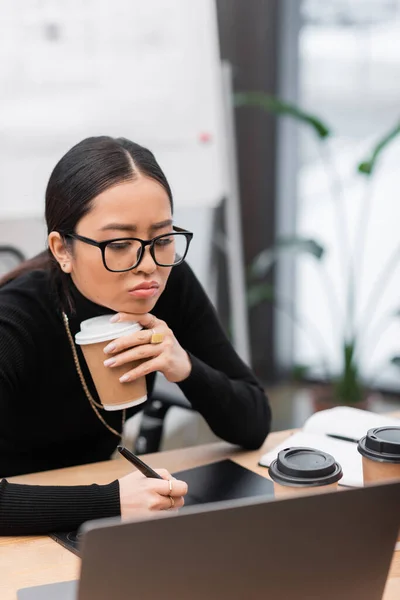 Bored asian designer holding coffee to go and using graphic tablet in office — Stock Photo