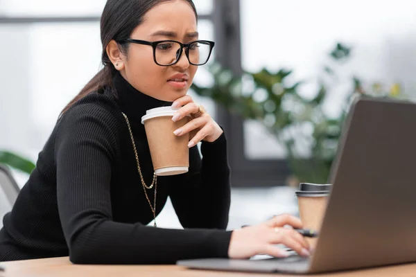 Upset asian designer holding paper cup and using laptop in office — Stockfoto