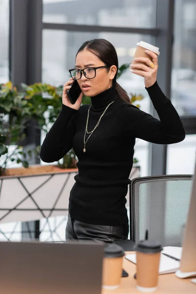 Злой азиатский дизайнер разговаривает на смартфоне и держит кофе, чтобы пойти в студию — стоковое фото