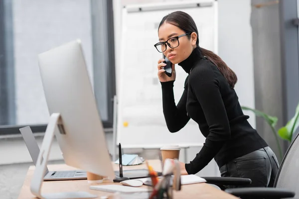 Asian designer in eyeglasses looking at computer and talking on smartphone in studio — Foto stock