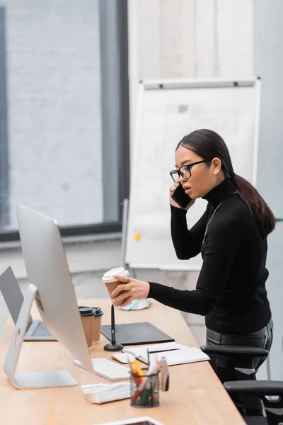 Young asian designer talking on smartphone and holding coffee near computer on table in office — Foto stock