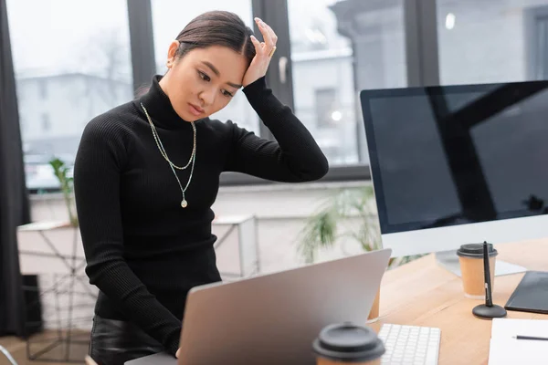 Worried asian interior designer using laptop near paper cups in office — Stockfoto