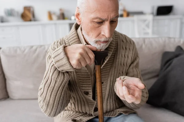 Senior man in knitted cardigan sitting with walking cane and looking at trembling hand while suffering from parkinson disease — Stock Photo