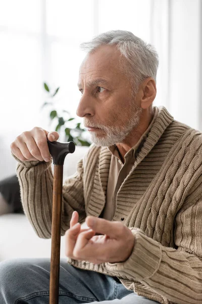 Depressed man with parkinsonian syndrome sitting with walking cane and looking away at home — Stockfoto