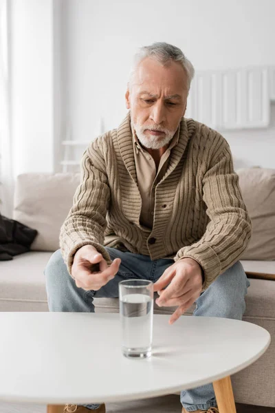 Старший седой мужчина с болезнью Паркинсона и дрожью в руках сидит возле стакана воды на столе дома — стоковое фото
