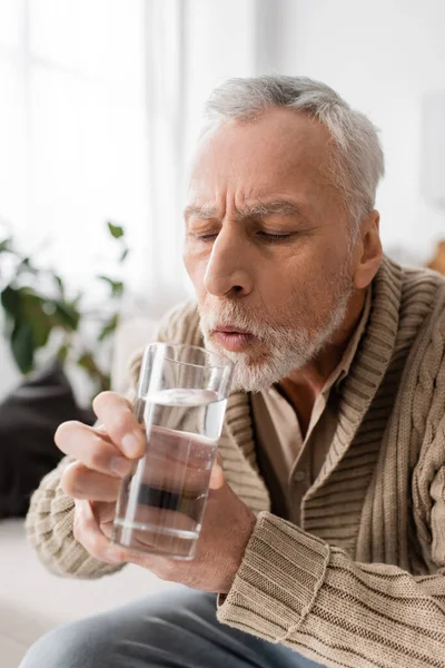 Седой мужчина с паркинсонизмом держа стакан воды в дрожащих руках дома — стоковое фото