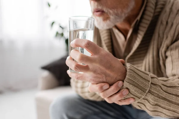 Cropped view of aged man with parkinsonism holding glass of water in trembling hands while sitting at home — Foto stock
