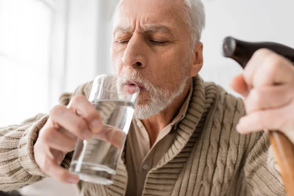 Пожилой человек с болезнью Паркинсона и дрожащими руками держа стакан воды и трость на размытом переднем плане — стоковое фото