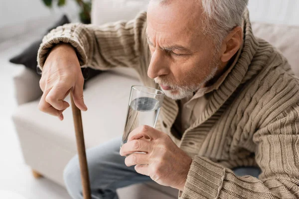 Літній чоловік з хворобою Паркінсона тримає склянку води в тремтячій руці, сидячи з ходячою тростиною вдома — стокове фото