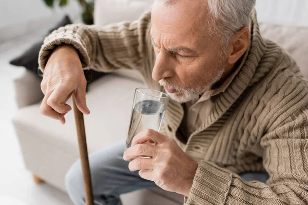 Aged man with parkinson disease holding glass of water in trembling hand while sitting with walking cane at home — Stock Photo