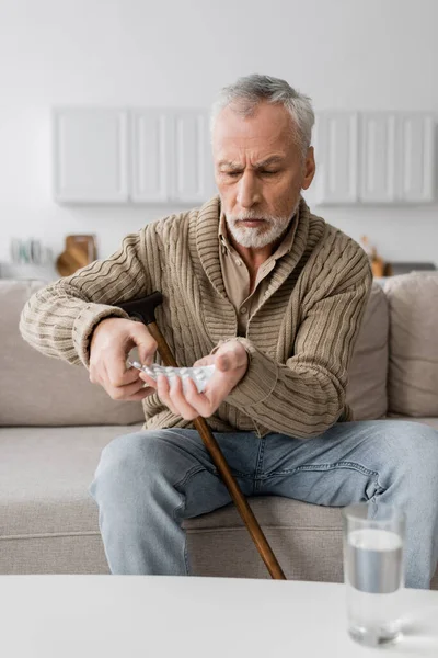 Aged man with parkinson syndrome holding pills in trembling hands while sitting on couch at home — Stockfoto