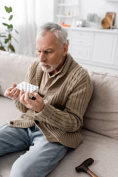 Пожилой человек с болезнью Паркинсона сидит на диване в вязаном кардигане и держит таблетки в дрожащих руках — стоковое фото