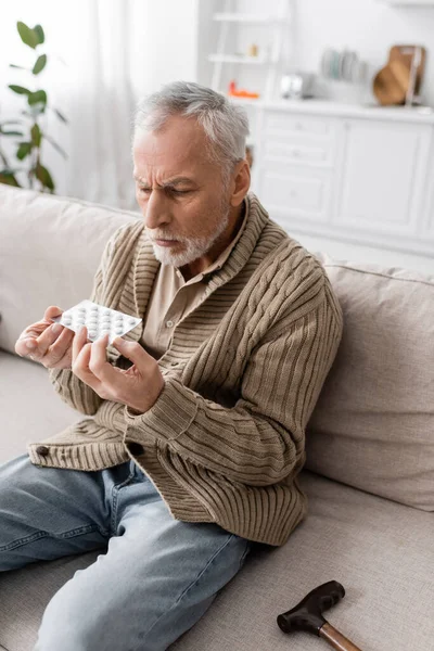 Senior man with parkinson disease sitting on couch in knitted cardigan and holding pills in trembling hands — Stock Photo