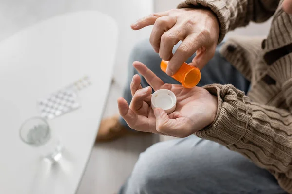Partial view of man with parkinson disease and tremor in hands holding pills container near blurred glass of water — Stockfoto
