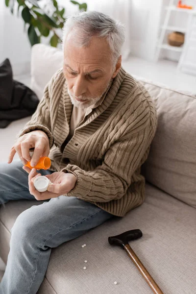 Grey haired man suffering from parkinsonism and hands tremor holding pills container near walking cane on couch at home — Stock Photo