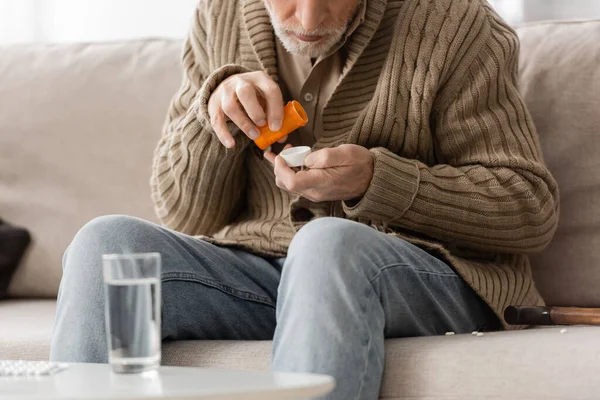 Partial view of aged man with parkinson syndrome sitting near blurred glass of water and holding pills container in trembling hands — Foto stock