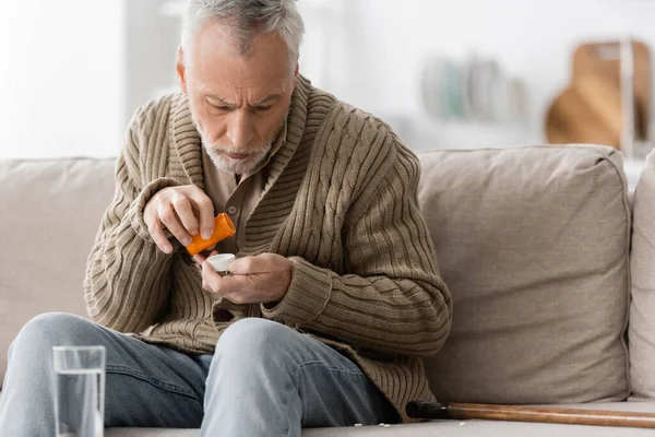 Седой мужчина с синдромом Паркинсона и дрожью рук сидит с контейнером для таблеток рядом с тростью и стаканом воды — стоковое фото