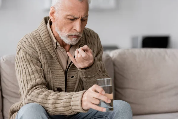 Senior man with parkinsonian syndrome sitting on couch and holding pill and glass of water in trembling hands — Stock Photo