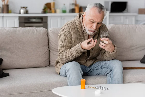 Senior man with parkinsonian syndrome holding pill and glass of water while sitting on couch near medication on table — Stock Photo