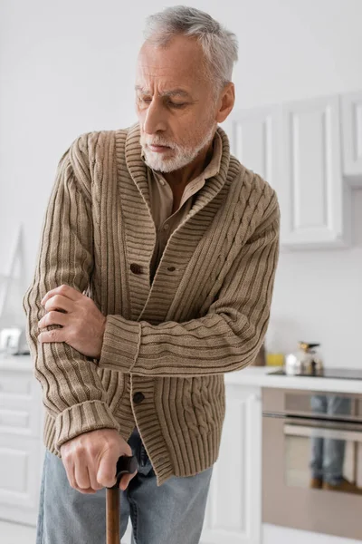Tensed man in knitted cardigan standing with walking cane in kitchen while suffering from parkinson disease — Stock Photo