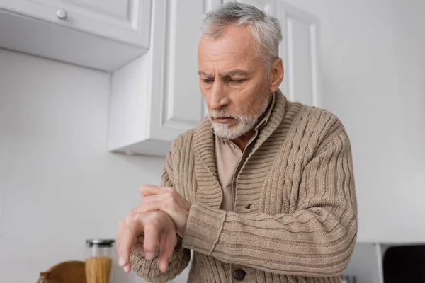 Седой мужчина с болезнью Паркинсона смотрит на дрожащие руки на кухне — стоковое фото