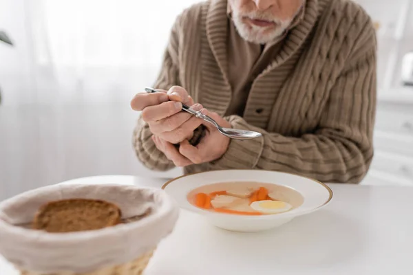 Частичный вид пожилого человека с синдромом Паркинсона, сидящего с ложкой в дрожащих руках возле супа и хлеба на кухне — стоковое фото