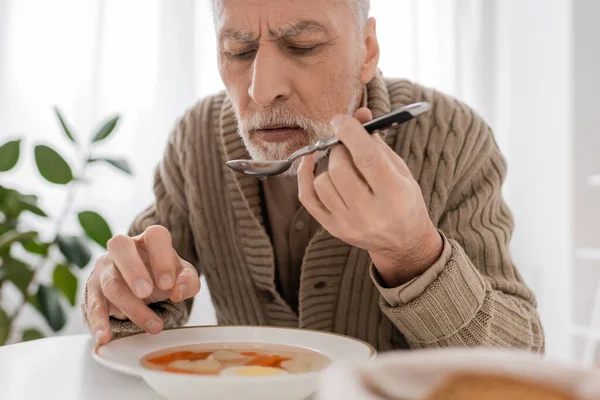 Senior bearded man suffering from parkinsonism and holding spoon in trembling hand near plate with soup in kitchen - foto de stock