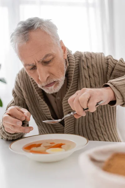 Aged man with parkinson disease and hands tremor sitting with spoon near plate with soup in kitchen — Stock Photo
