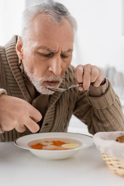 Седой мужчина с синдромом Паркинсона ест суп на ужин на кухне — стоковое фото