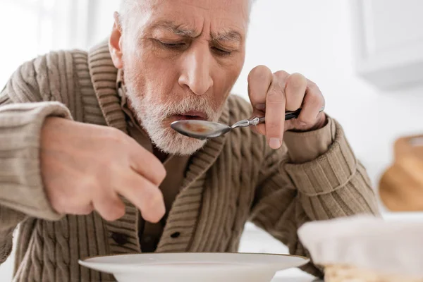 Пожилой мужчина, страдающий синдромом Паркинсона, держа ложку в дрожащей руке во время ужина дома — стоковое фото