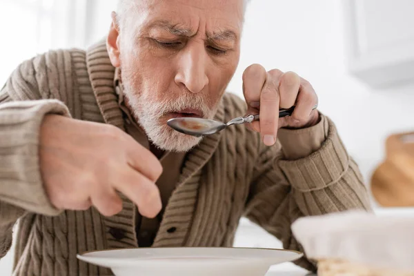 Aged man suffering from parkinsonian syndrome holding spoon in trembling hand while having dinner at home — Stock Photo