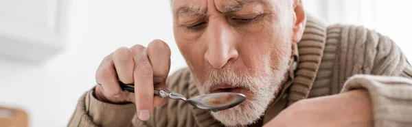 Bearded senior man with parkinsonian syndrome and hands tremor holding spoon while having dinner at home, banner — Stockfoto