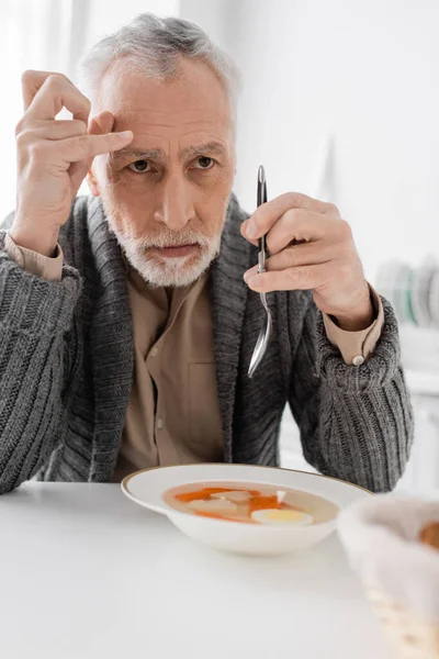 Depressed man with parkinsonian syndrome looking away while holding spoon near soup in kitchen — Stock Photo