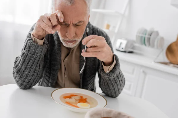 Depressed man with parkinson syndrome holding spoon while sitting with closed eyes near plate with soup in kitchen — Stock Photo