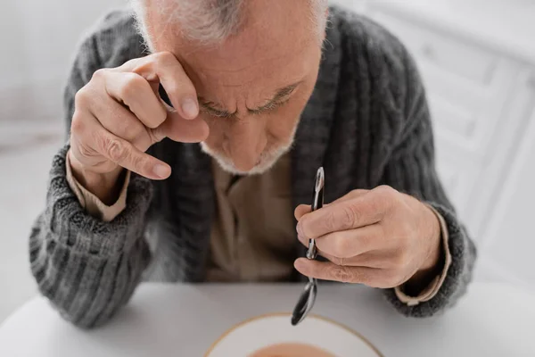 Senior man suffering from parkinson disease and hands tremor sitting with spoon during lunch in kitchen — Stock Photo