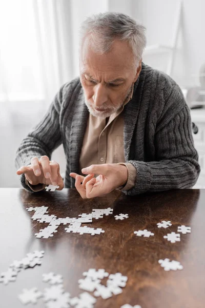 Aged man with parkinsonian syndrome and tremor in hands combining jigsaw puzzle on table at home — Stock Photo